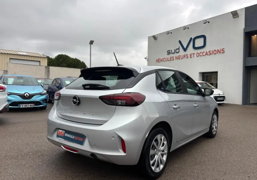 Opel Corsa gris clair vue 3/4 arrière droit, avec jantes classiques et feux arrière foncés, stationnée devant un concessionnaire.