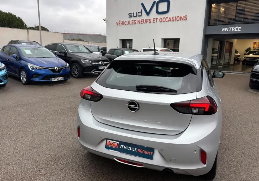 Vue 3/4 arrière d’une Opel Corsa gris clair 2024 garée devant un showroom avec feux arrière allumés.