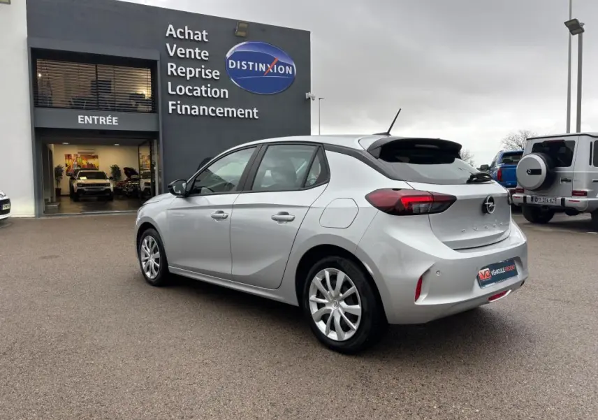 Opel Corsa gris clair vue 3/4 arrière droit, avec jantes classiques 16 pouces, stationnée devant un garage.