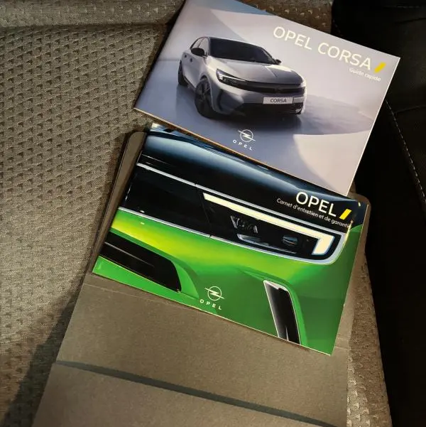 Deux brochures Opel Corsa posées sur un siège en tissu gris, l'une montrant une Corsa grise de face, l'autre un gros plan sur un phare avant vert.