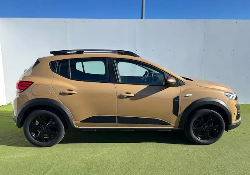 Profil droit d'une Dacia Sandero III Stepway beige Safari avec jantes noires et barres de toit gris Mégalithe.