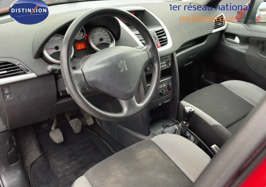 Vue intérieure côté conducteur de la Peugeot 207 rouge métal, montrant le volant, tableau de bord et sièges tissu noir et gris.