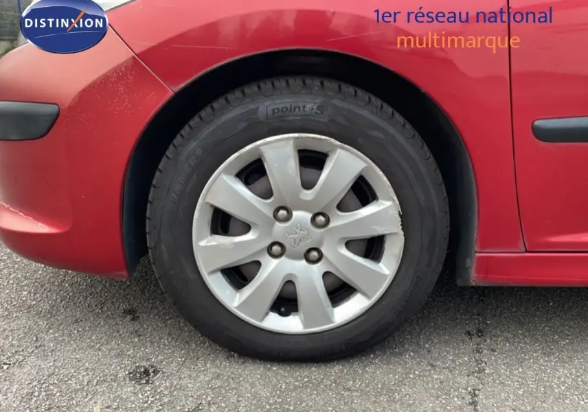 Gros plan sur la roue avant gauche d'une Peugeot 207 rouge métal, mettant en valeur la jante et la carrosserie.