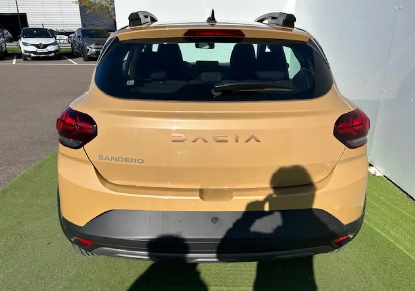 Vue arrière d'une Dacia Sandero beige Safari 2025 avec logo et feux arrière LED, sur sol vert artificiel.