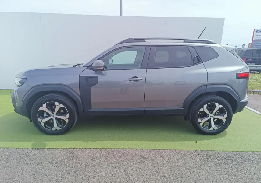 Vue latérale gauche du Dacia Duster gris schiste métal 2026 avec jantes alliage 18 pouces et barres de toit noires.