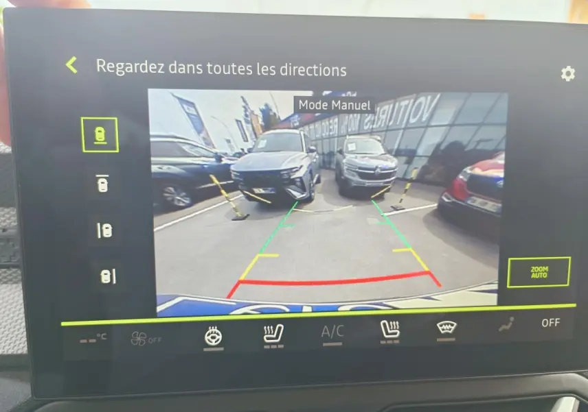 Vue caméra de recul montrant un Dacia Duster gris schiste en 3/4 avant gauche sur un parking devant un bâtiment vitré.