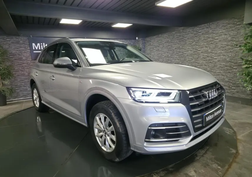 Audi Q5 gris métallisé en 3/4 avant droit, avec calandre noire et phares LED allumés dans un showroom.