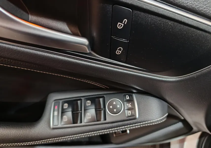 Gros plan sur la commande des vitres et rétroviseurs côté gauche en cuir noir avec surpiqûres blanches dans une Mercedes GLA gris clair.