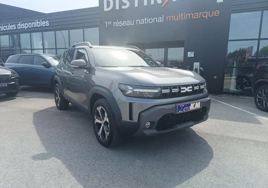 Dacia Duster III gris schiste métal en 3/4 avant droit, avec jantes alliage et barres de toit noires visibles.