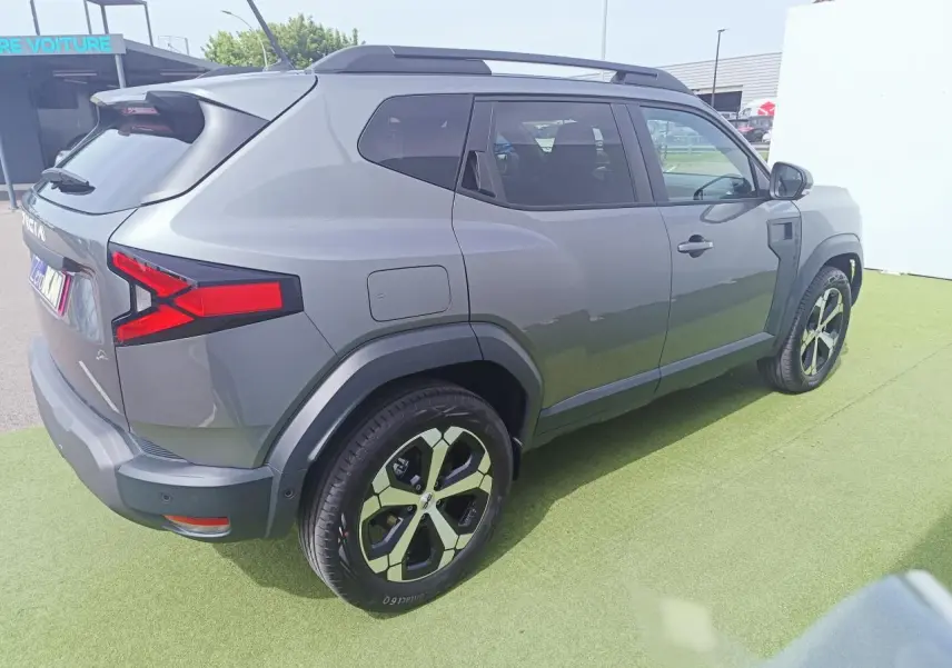Vue 3/4 arrière droite du Dacia Duster 2026 gris schiste métal avec jantes alliage 18 pouces et barres de toit noires.