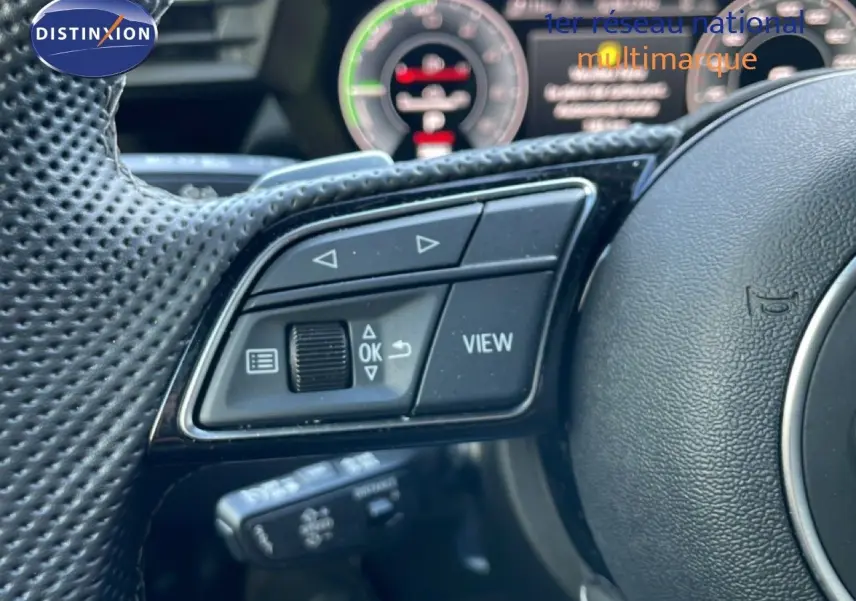 Gros plan sur les commandes gauche du volant perforé d'une Audi A3 Sportback 2025, avec affichage numérique flou en arrière-plan.