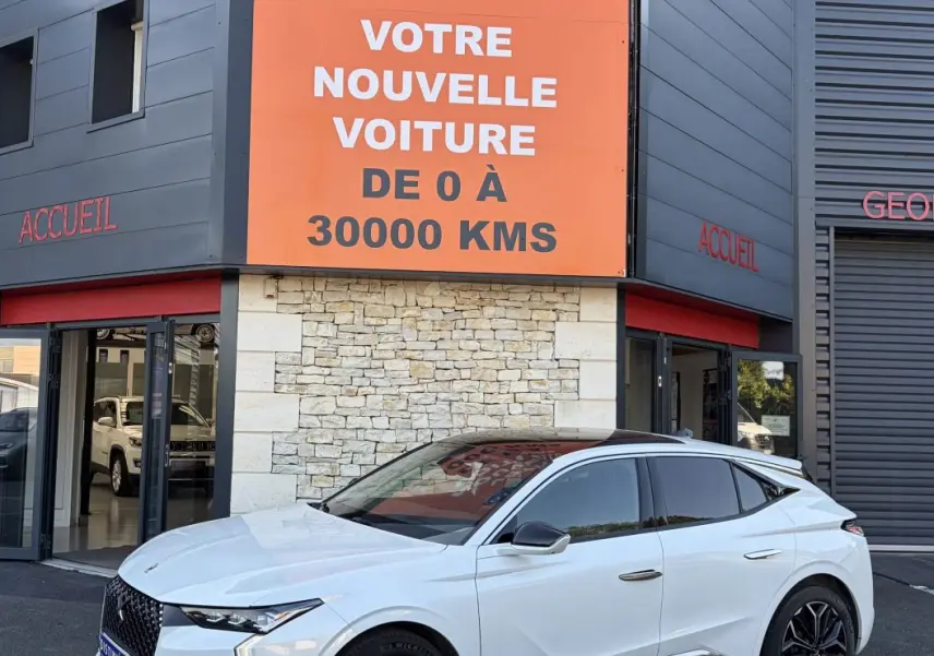 DS4 blanc vue 3/4 avant droit, jantes noires, calandre distinctive, devant un bâtiment avec panneau orange Distinxion.