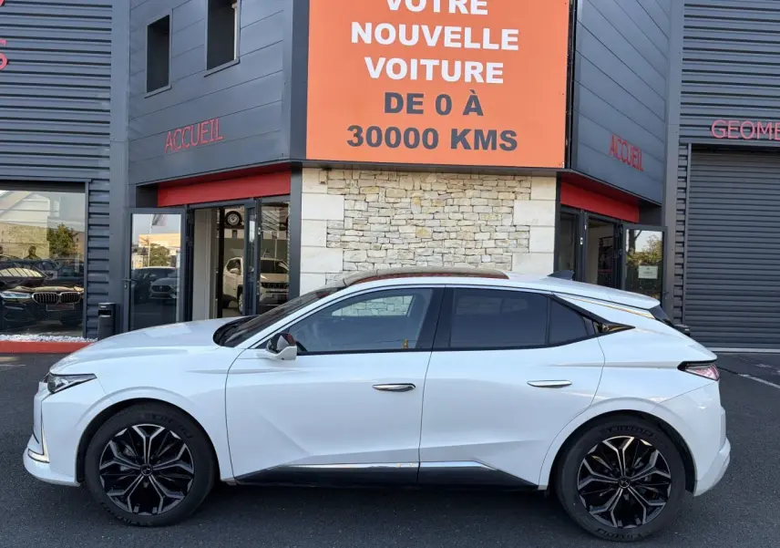 Vue latérale gauche d'une DS4 blanche 2024 avec jantes noires et vitres teintées, devant un bâtiment commercial.