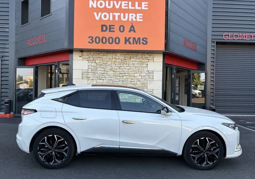 Vue de profil côté gauche d’une DS4 blanche 2024 avec jantes noires et toit panoramique, devant un bâtiment commercial.