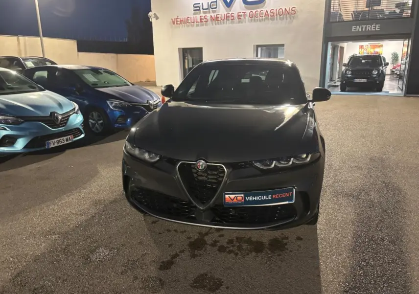 Vue avant d'une Alfa Romeo Tonale gris foncé garée de nuit devant un concessionnaire, avec phares allumés.