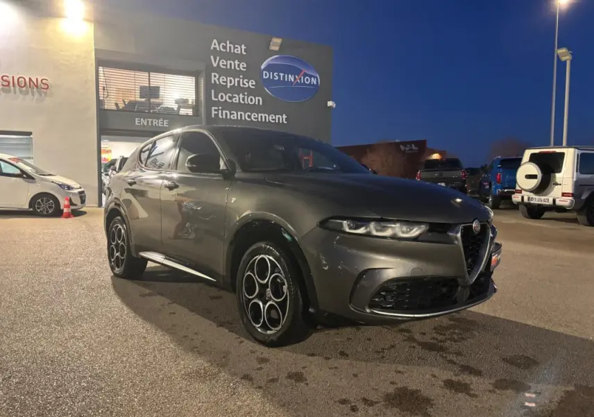Vue 3/4 avant gauche d'une Alfa Romeo Tonale gris métallisé avec jantes alliage 18 pouces, stationnée de nuit devant un showroom.
