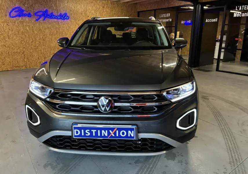 Vue avant d'un Volkswagen T-Roc gris 2023 avec éclairage LED allumé et calandre chromée dans un showroom.