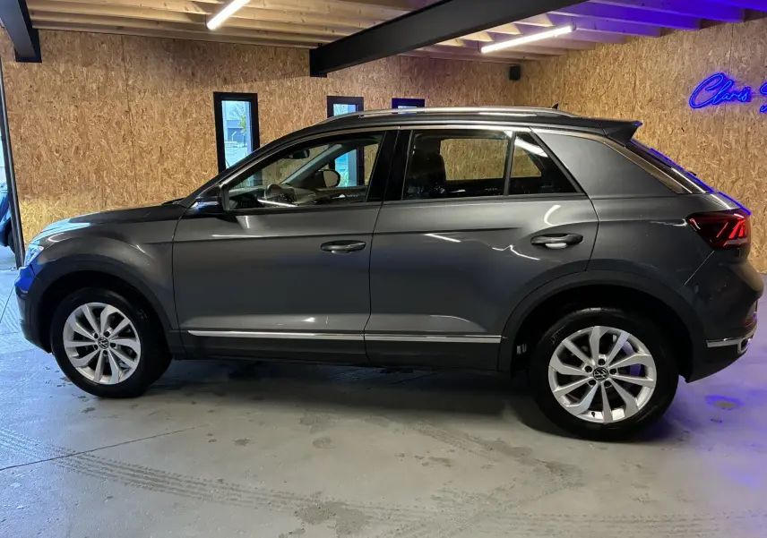 Vue de profil côté gauche d’un Volkswagen T-Roc gris 2023, SUV compact avec jantes alliage et toit noir en intérieur showroom.