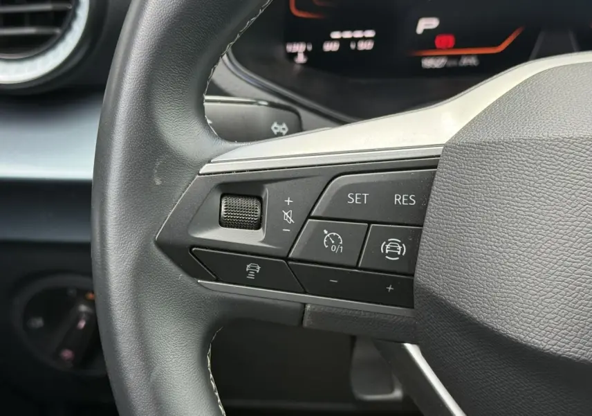 Gros plan sur les commandes de régulateur de vitesse au volant noir d’un SEAT Arona 2025, tableau de bord digital en arrière-plan.