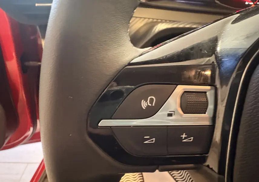 Gros plan sur les commandes vocales et volume au volant d’une Peugeot 208 Active rouge, côté gauche, intérieur 2024.