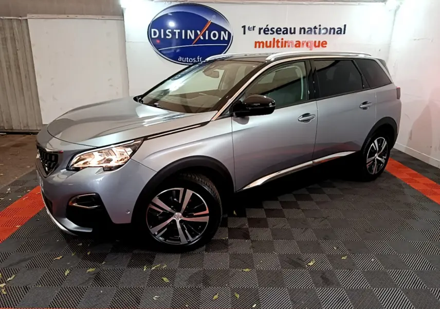 Peugeot 5008 gris clair vue 3/4 avant droit, avec jantes alliage et barres de toit chromées en intérieur showroom.