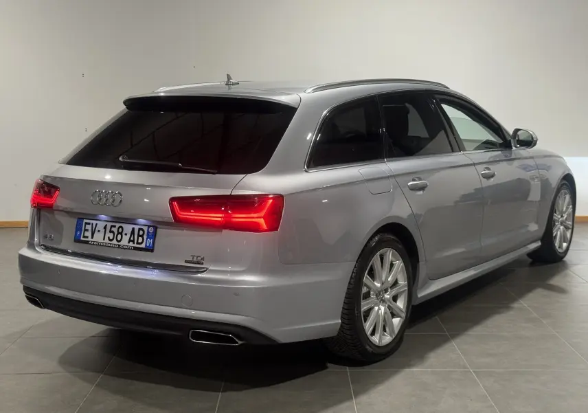 Audi A6 Avant gris vue 3/4 arrière droit avec feux allumés et jantes alliage, en intérieur.
