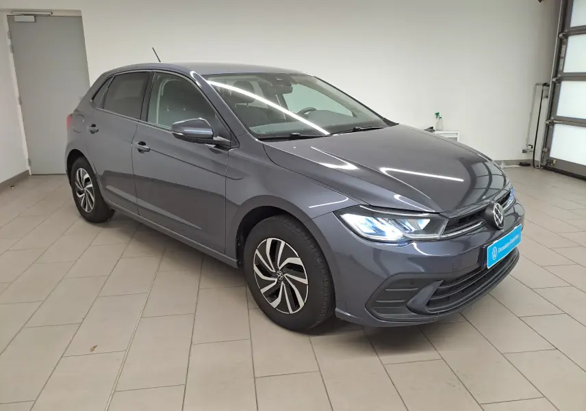 Volkswagen Polo gris cendre vue 3/4 avant droit en intérieur avec phares allumés et jantes distinctives.