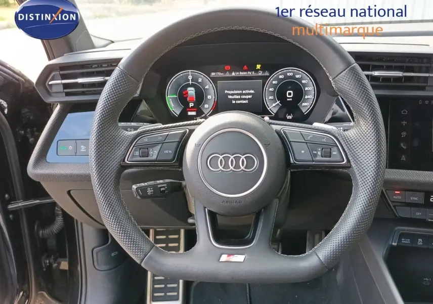 Vue intérieure centrée sur le volant perforé d’une Audi A3 Sportback noire, avec tableau de bord numérique et commandes intégrées.