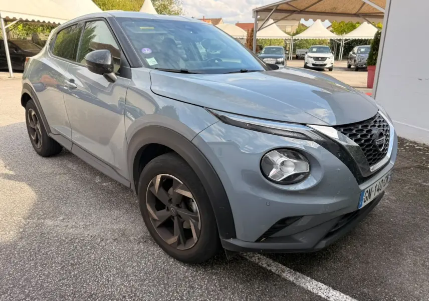 Nissan Juke gris clair vu en 3/4 avant droit, avec jantes noires et calandre distinctive en V chromé.