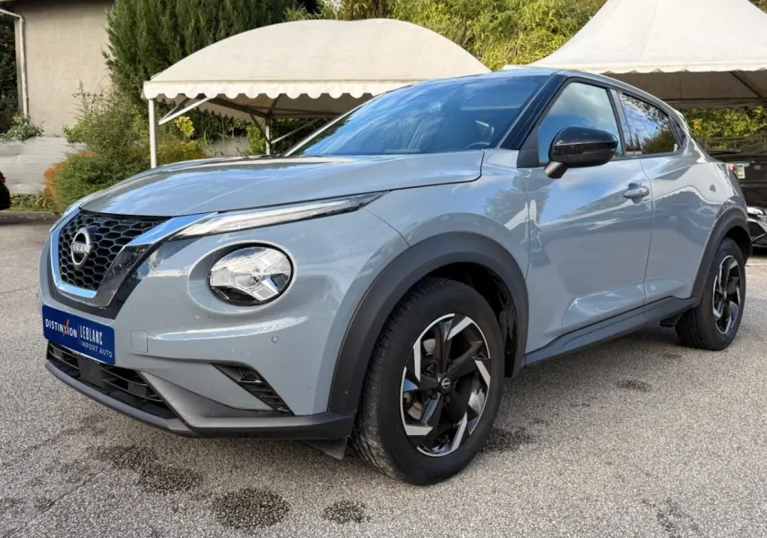 Nissan Juke gris clair vu de 3/4 avant droit, avec phares ronds et jantes noires à motifs géométriques.