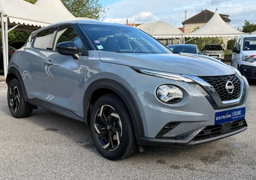 Nissan Juke gris clair vue 3/4 avant droit, avec calandre noire en V et jantes noires à motifs argentés.
