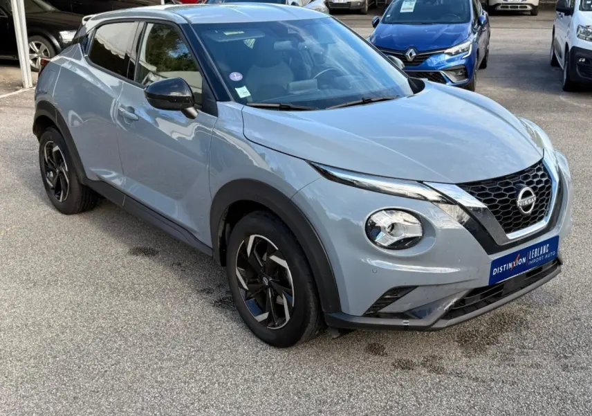 Nissan Juke gris clair vu en 3/4 avant droit, avec calandre noire et jantes alliage noires distinctives.