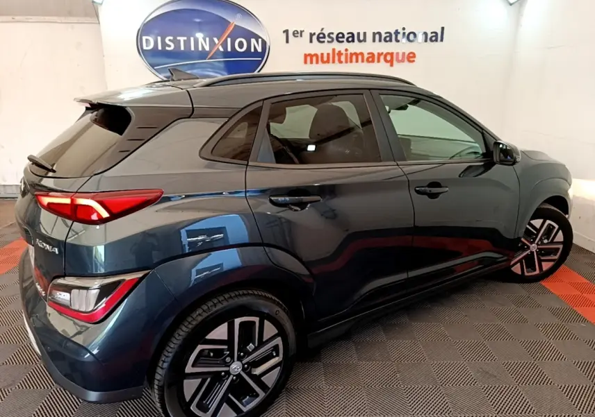 Vue 3/4 arrière droite d’un Hyundai Kona électrique bleu 2023 avec feux LED et jantes distinctives en intérieur showroom.
