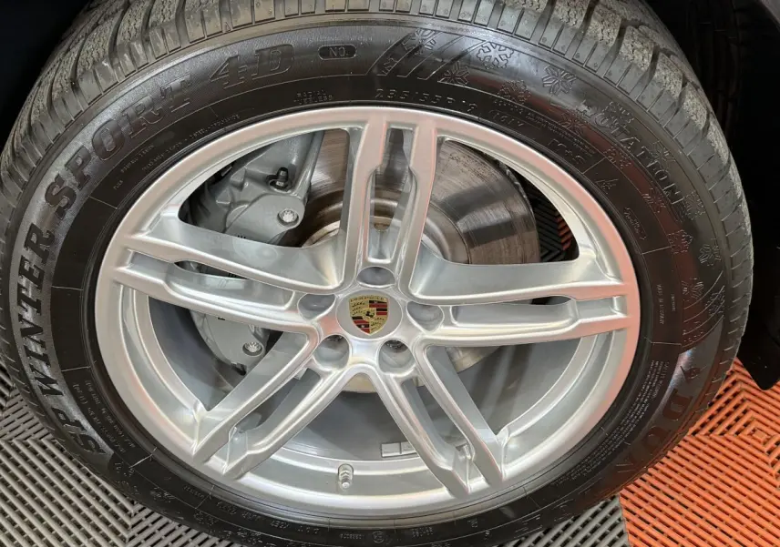 Gros plan sur la jante alu argentée avec logo Porsche centrale et pneu hiver Dunlop sur Porsche Macan gris foncé.