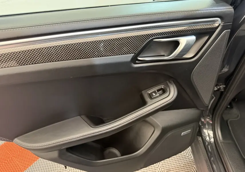 Vue intérieure côté gauche de la porte arrière d’un Porsche Macan 3.0 S gris foncé avec garniture carbone et bouton de lève-vitre.