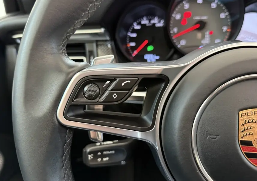 Gros plan sur le volant en cuir et alu du Porsche Macan 3.0 S, avec commandes multifonctions et tableau de bord partiellement visible.