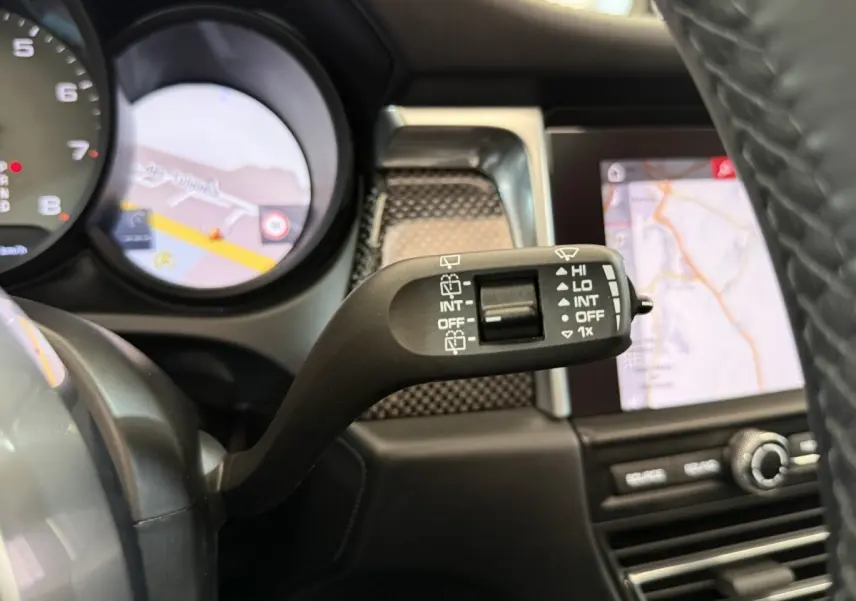 Gros plan sur la commande d’essuie-glace à gauche du volant en cuir du Porsche Macan 3.0 S gris foncé, avec écran tactile en arrière-plan.