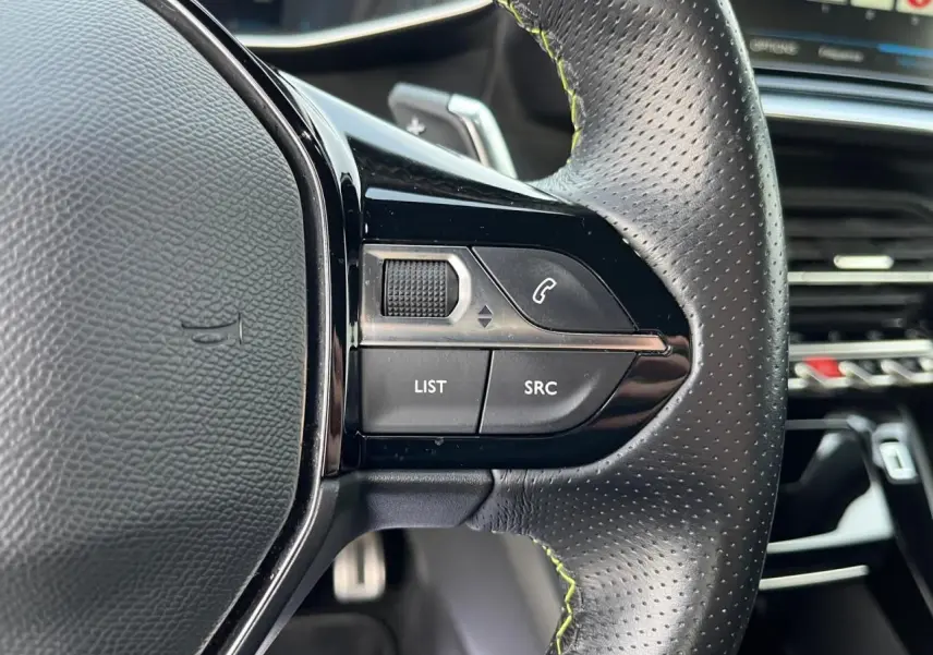 Gros plan sur les commandes gauche du volant en cuir perforé du Peugeot 2008 II gris Artense avec surpiqûres vertes.