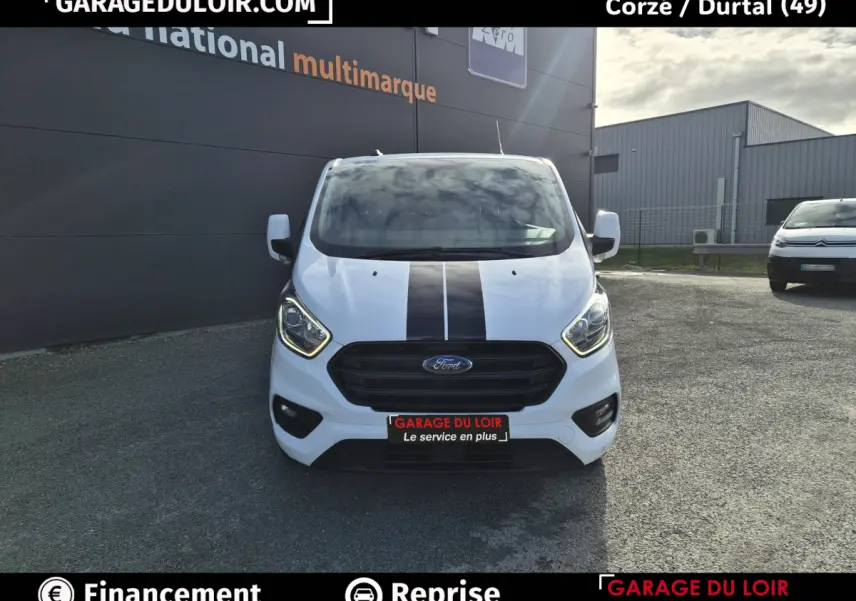 Vue avant d'un Ford Transit Custom blanc 2023 avec bande noire centrale et calandre noire, stationné devant un bâtiment.