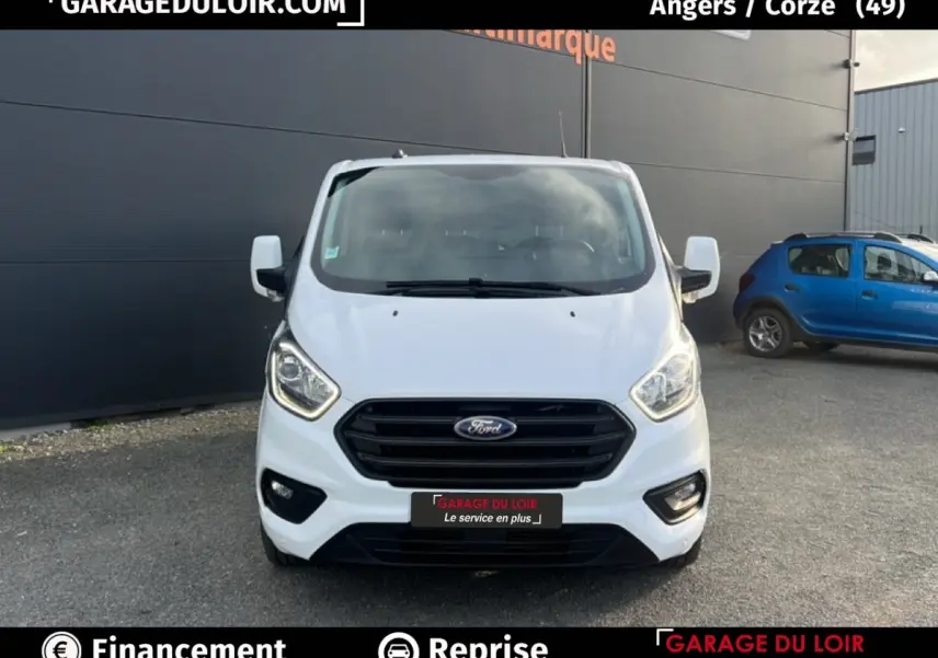 Vue de face d'un Ford Transit Custom Fourgon blanc 2023 avec calandre noire et feux allumés sur parking extérieur.