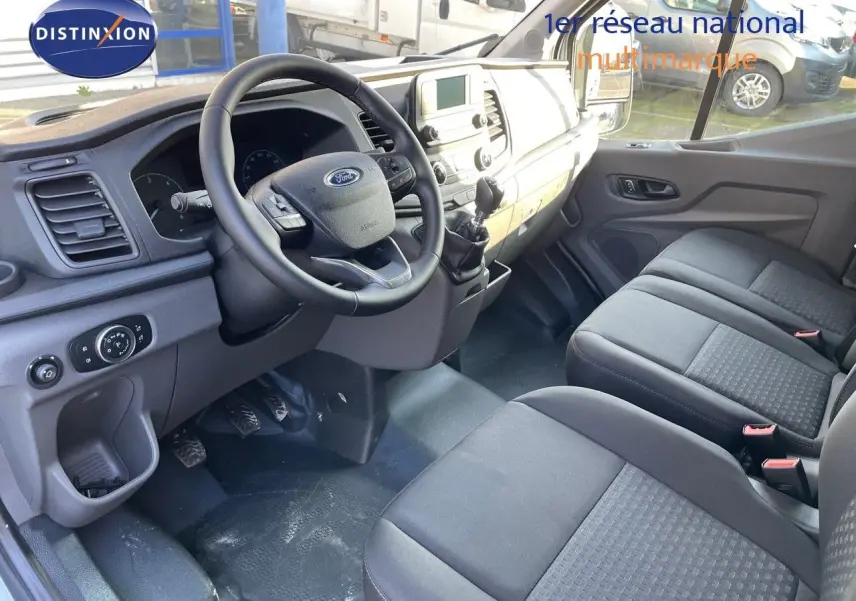 Vue intérieure côté conducteur du Ford Transit Fourgon 2023, montrant le volant, la boîte manuelle et les sièges gris.