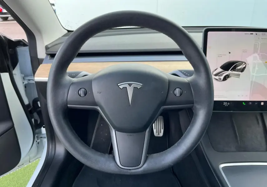 Vue centrée sur le volant noir d'une Tesla Model 3 blanche, avec écran tactile et intérieur partiellement en bois clair.