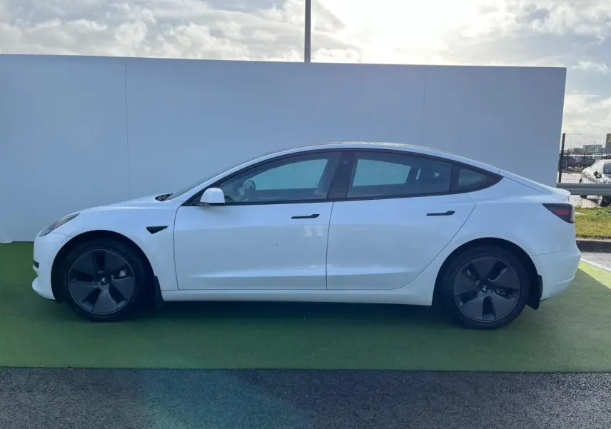 Profil côté gauche d'une Tesla Model 3 blanche 2022 avec jantes noires et poignées affleurantes ton carrosserie.
