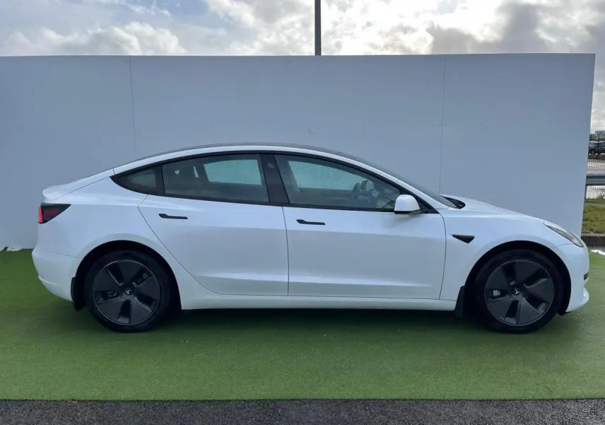 Profil côté gauche d'une Tesla Model 3 blanche 2022 avec jantes noires et poignées affleurantes ton carrosserie.