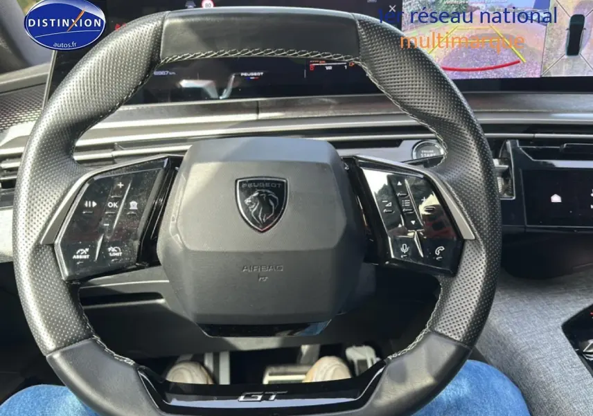 Vue rapprochée du volant cuir perforé noir avec logo Peugeot et inscription GT, tableau de bord numérique en arrière-plan.