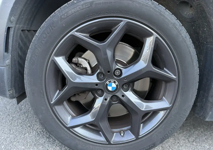 Gros plan sur la roue avant gauche d’un BMW X1 blanc, jante alliage gris foncé à cinq branches doubles.