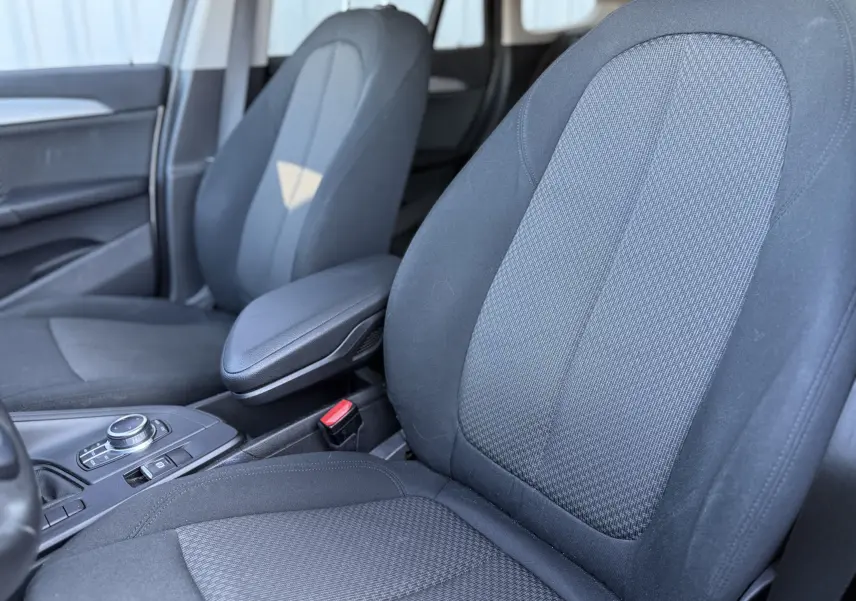 Gros plan sur les sièges avant en tissu noir de la BMW X1 20D 2018, avec accoudoir central et console de commandes visible.