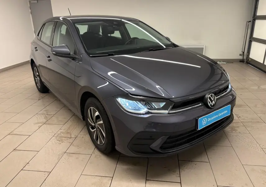 Volkswagen Polo gris cendre 2024 vue 3/4 avant droit dans un showroom avec phares allumés.