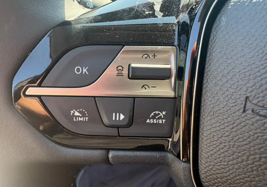 Gros plan sur les commandes de régulateur et limiteur de vitesse à gauche du volant d’une Peugeot 308 PHEV 2024 gris clair.