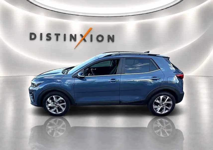Vue de profil côté gauche d'un KIA Stonic gris 2025 dans un showroom avec jantes alliage et barres de toit.